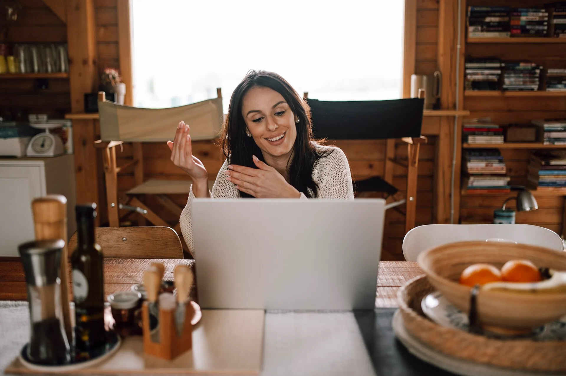 A woman is sitting at a wooden table with a laptop. She is smiling while talking on a video call. The room has wooden walls and a bright window. There are kitchen items on the table. Virtual assistant agencies: what to check before hiring