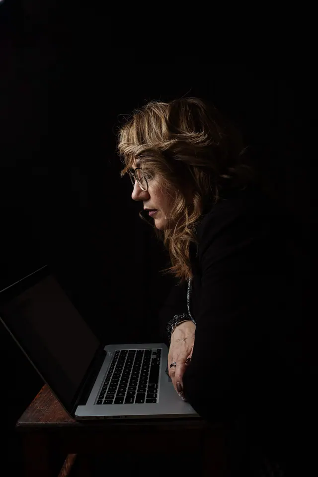 A focused middle aged woman uses a laptop while working late at night in a low-light apartment interior. The image illustrates remote work, concentration, digital communication and. CRA contractor rules for a virtual assistant agency: the factors that decide