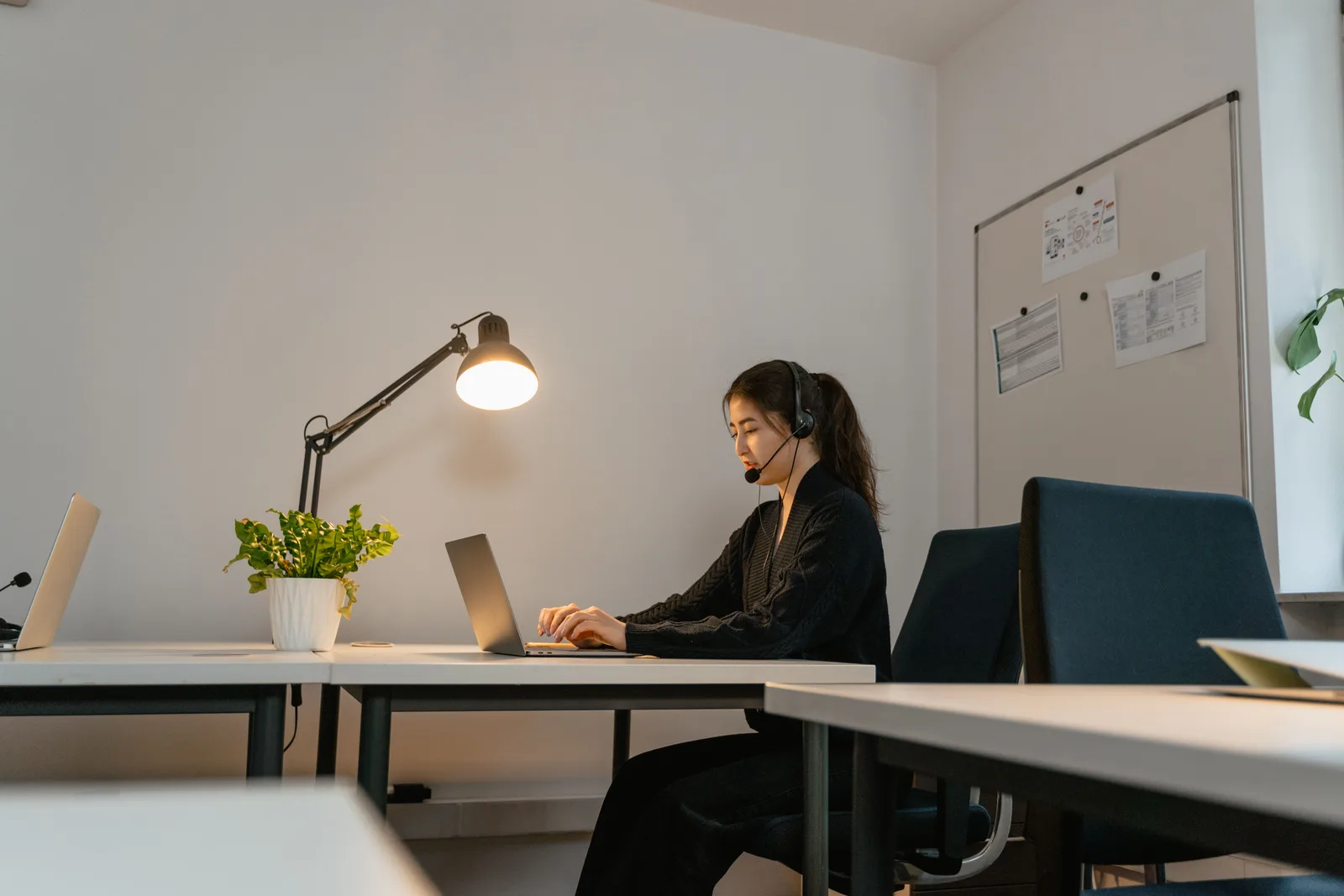 A woman in a modern office setting, wearing a headset and working on a laptop under a desk lamp. 7 points worth checking on virtual assistant agency startup