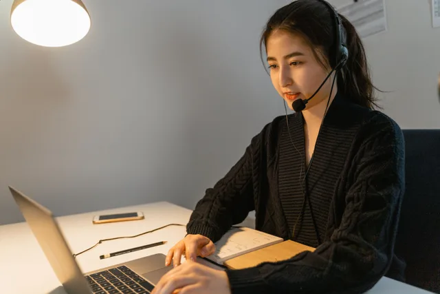 Asian woman using a laptop with a headset in a modern office setting. 7 things worth knowing about virtual assistant agency trends